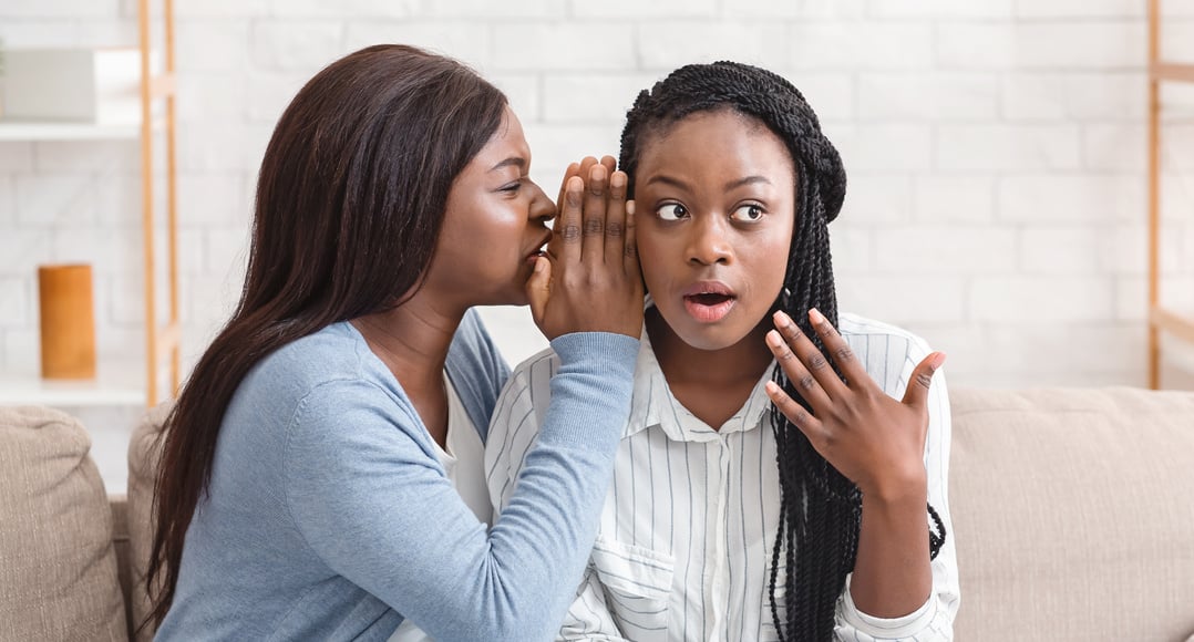 Young black girl whispering secret to her shocked friend's ear